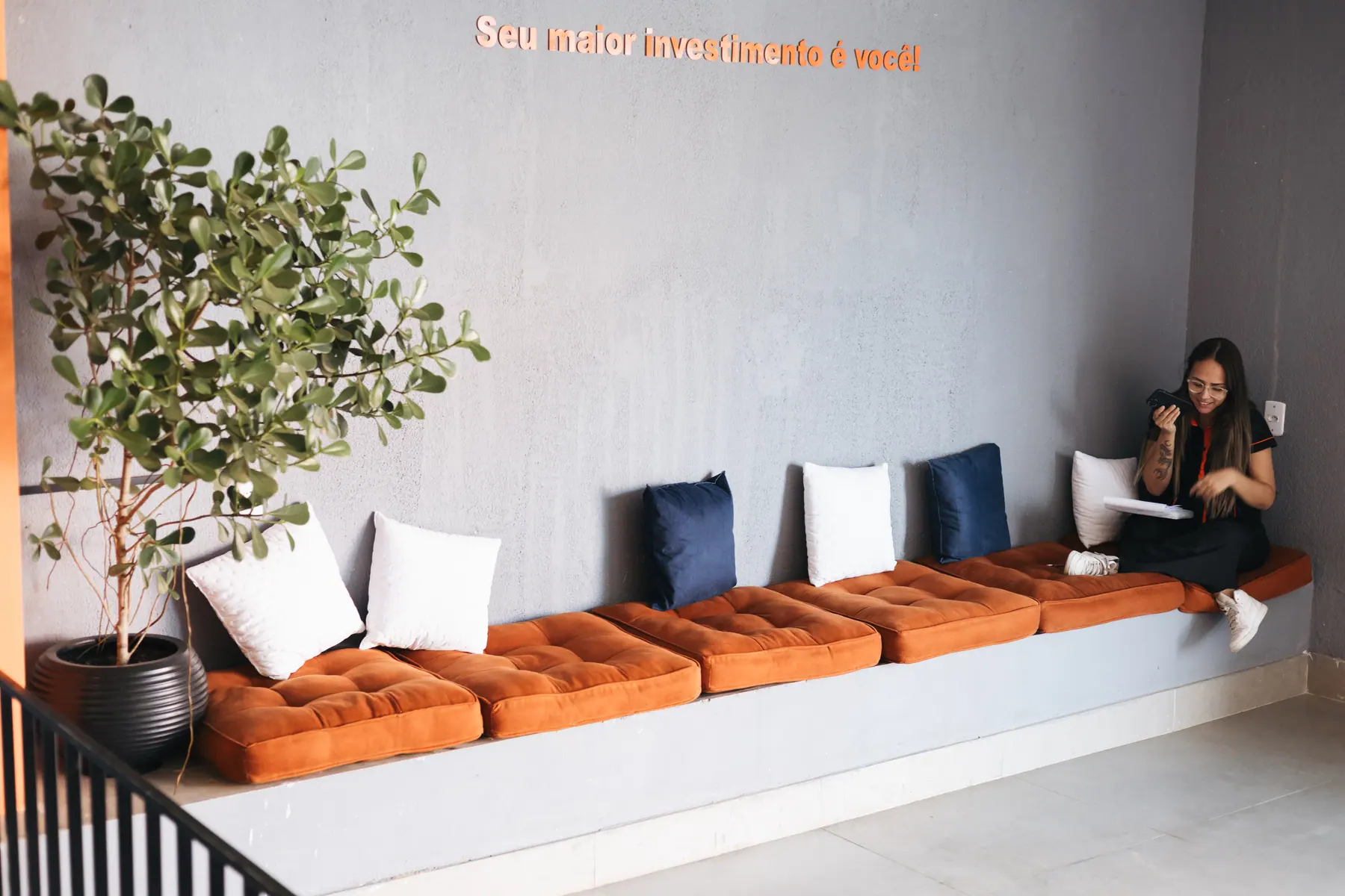 Lounge com bancos estofados laranja, almofadas e planta, onde uma aluna descansa
